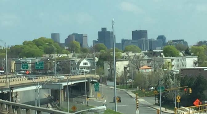 An atmospheric view from an expressway with a city skyline, street layouts, and tall buildings visible in the background.