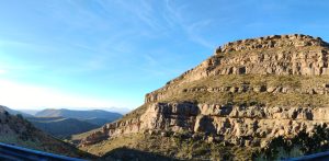 A wide landscape photograph capturing a massive, rugged rock mountain rising into the sky, overlooking a vast valley filled with a rolling sea of smaller mountain peaks below in New Mexico.