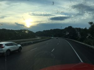 A view from inside a truck looking through a weathered windshield at a curving mountain highway lined with evergreen trees under a dramatic sunset sky.