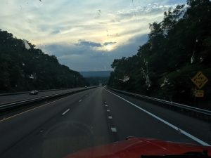 Spirit Presence A dramatic traveler’s photograph taken from a vehicle looking down an asphalt highway lined with dense green evergreen trees on both sides under a dark twilight sky, where a beautiful sunset is glowing softly behind a massive dark cloud with mountains in the background.