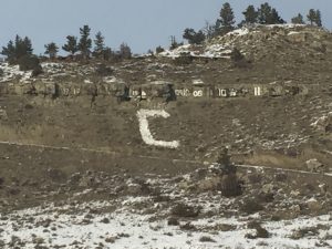 Close-up view of a rugged, rocky mountain featuring a large, distinct white block letter C built into the side, surrounded by scattered green trees and patches of fresh white snow under an overcast sky.