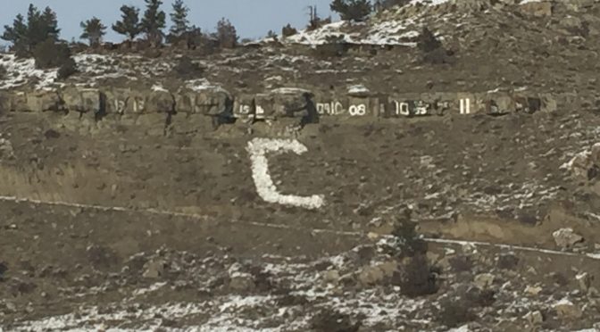 Close-up view of a rugged, rocky mountain featuring a large, distinct white block letter C built into the side, surrounded by scattered green trees and patches of fresh white snow under an overcast sky.