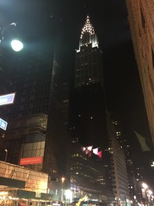 A striking architectural perspective looking up at a massive, towering high-rise building with many stories and brilliant glowing lights at the top, captured at night from near the Grand Hyatt in New York.