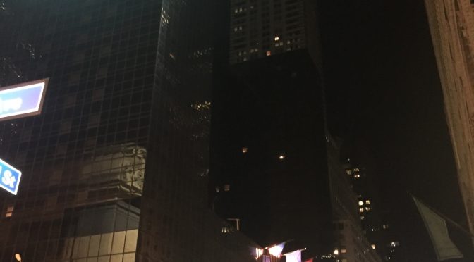 Spirit Presence visible in a striking architectural perspective of a massive, supertall skyscraper with countless stories rising very high into the New York night sky near the Grand Hyatt, featuring brilliant glowing lights illuminating the towering structure at the top and casting a steady, hopeful light far above the city streets.