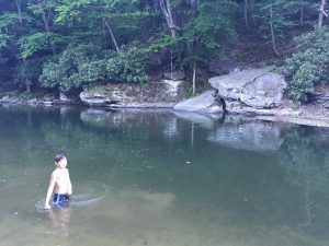  A young boy standing in clear, shallow, freezing water surrounded by smooth river stones, with a massive ridge of craggy boulders and steep, mountainous rock formations rising sharply along the opposite bank under a soft sky.